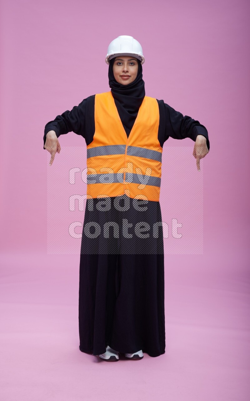 Saudi woman wearing Abaya with engineer vest and helmet standing interacting with the camera on pink background