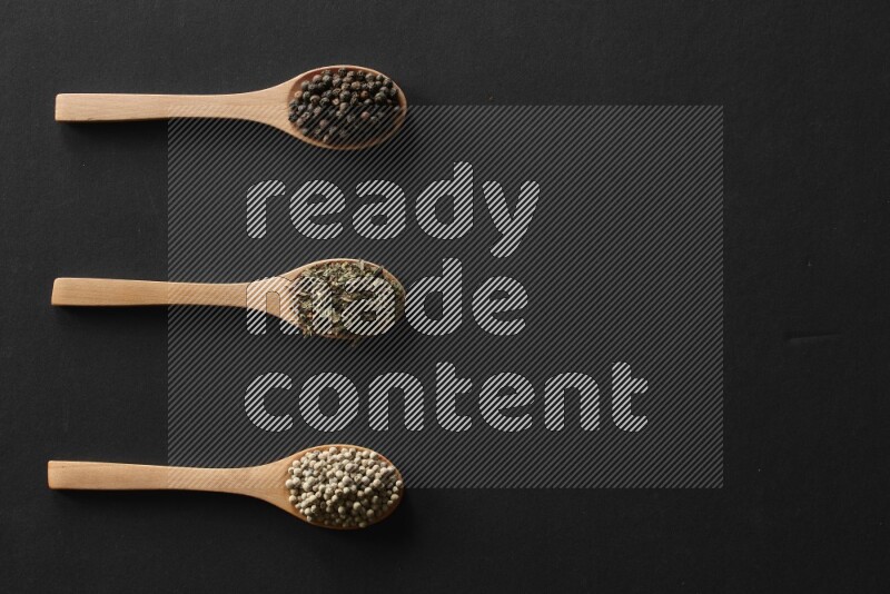 الفلفل الأبيض والقرنفل والهيل والملح والفلفل الأسود وريحان مجفف في معالق خشبية علي خلفية سوداء