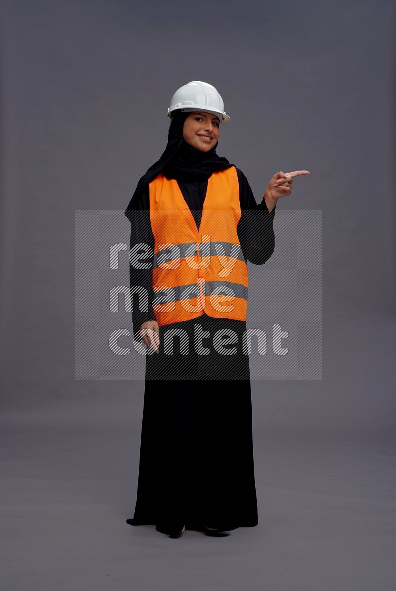 Saudi woman wearing Abaya with engineer vest standing interacting with the camera on gray background