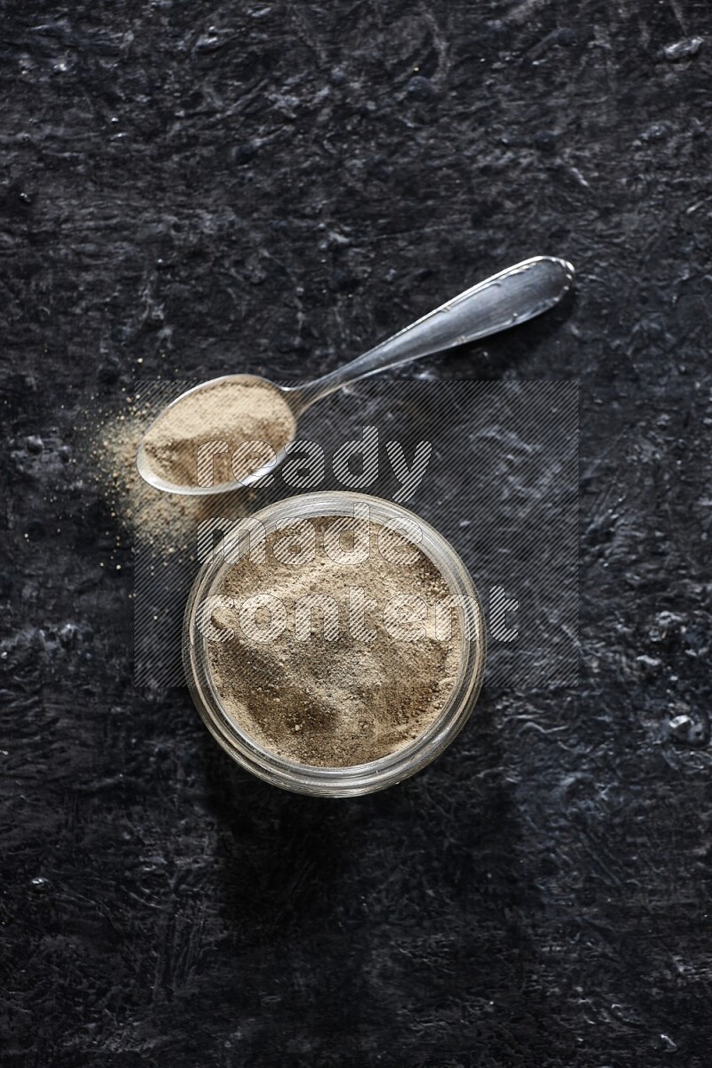 A glass jar and a metal spoon full of white pepper powder on textured black flooring