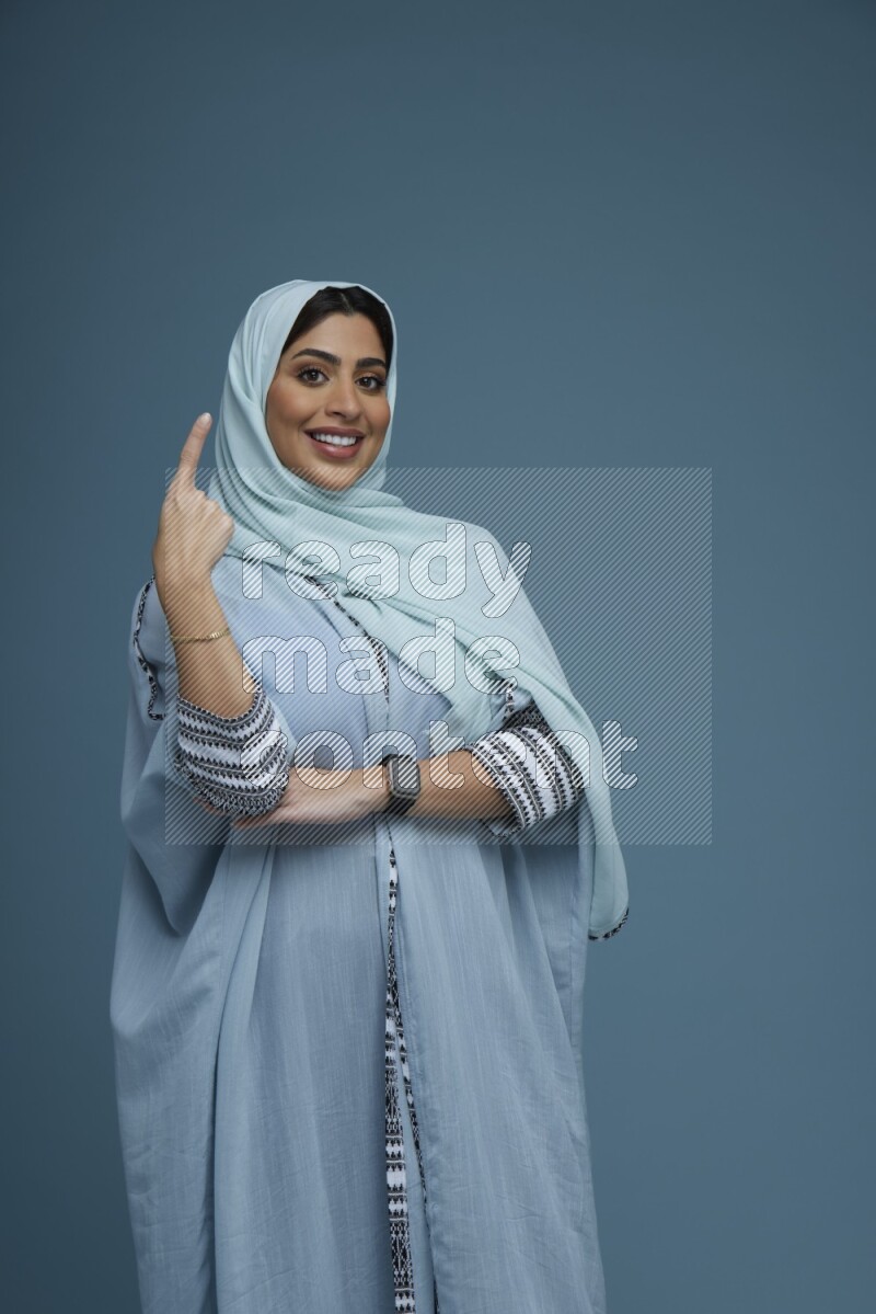 A Saudi woman pointing in a blue background wearing a blue Abaya with hijab