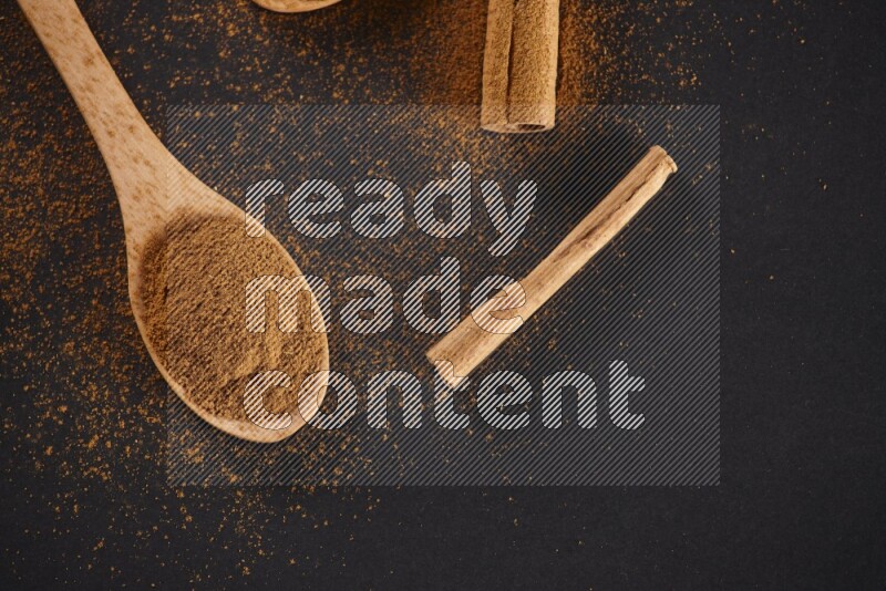Cinnamon powder in two wooden spoons with cinnamon sticks on black background