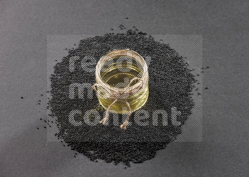 A glass jar full of black seeds oil surrounded by the seeds on a black flooring