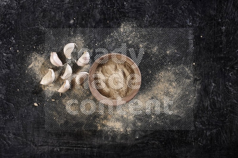 A wooden bowl full of garlic powder with some garlic cloves beside it on a textured black flooring