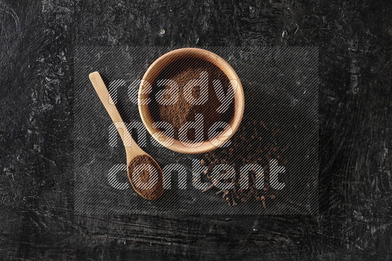 A wooden bowl and a wooden spoon full of cloves powder with spreaded cloves on a textured black flooring