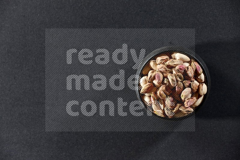 A black pottery bowl full of peeled pistachios on a black background in different angles