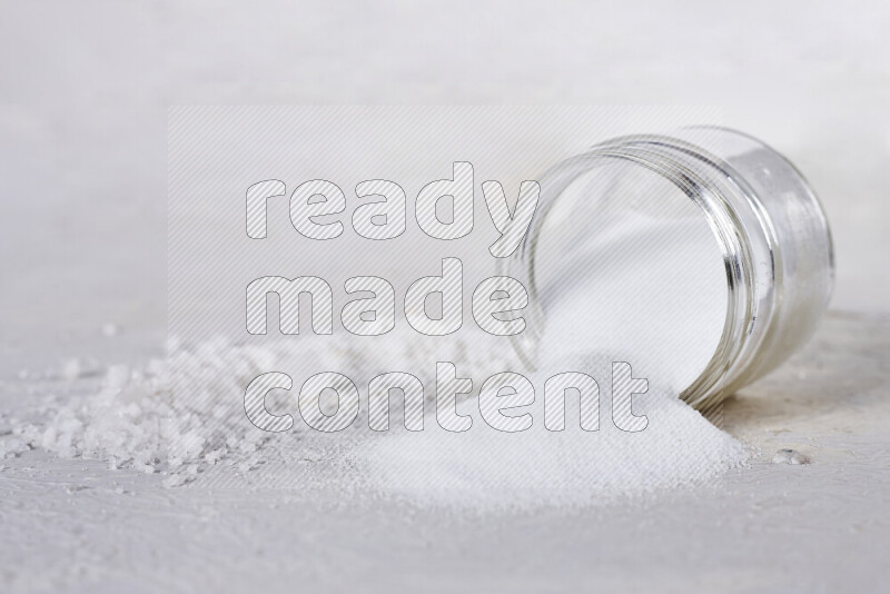 A glass jar full of table salt with some sea salt crystals beside it on a white background