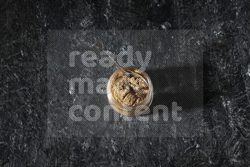 A glass jar and a metal spoon full of allspice powder on a textured black flooring