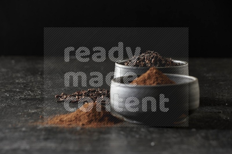 2 Black pottery bowls full of cloves and the other full of cloves powder on textured black flooring