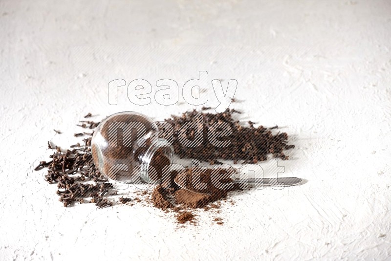 A flipped glass spice jar and a metal spoon full of cloves powder and powder came out of the jar with cloves spread on textured white flooring