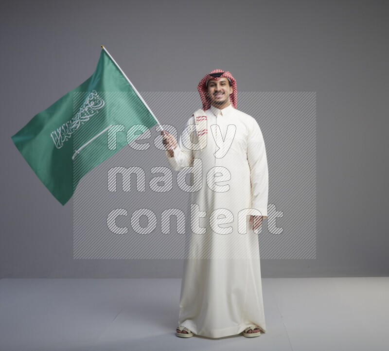 A Saudi man standing wearing thob and red shomag raising big Saudi flag on gray background