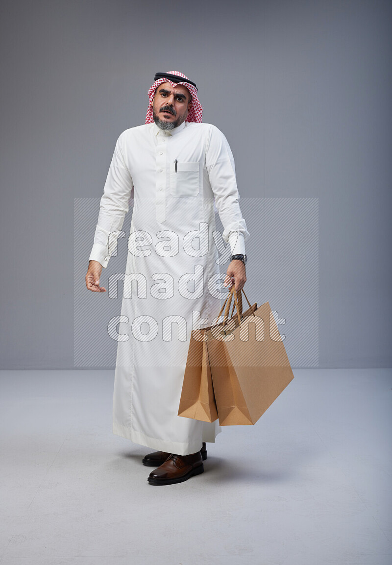 Saudi man Wearing Thob and red Shomag standing holding shopping bag on Gray background