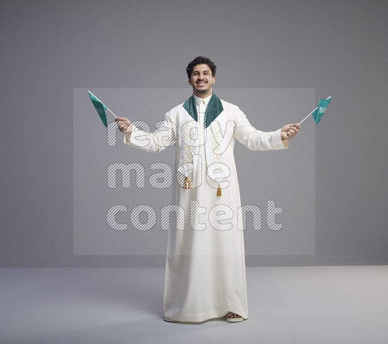 A Saudi man standing wearing thob and saudi flag scarf and holding small saudi flag on gray background