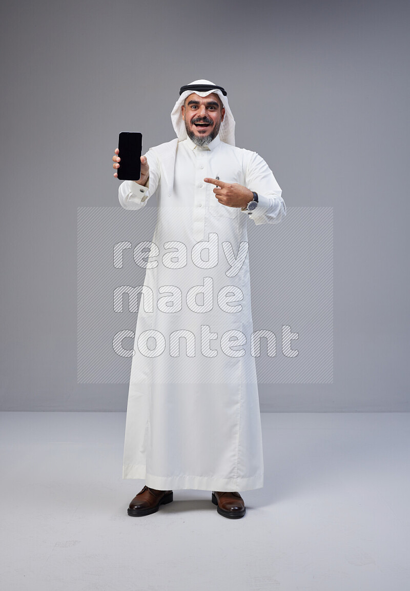 Saudi man Wearing Thob and white Shomag standing showing phone to camera on Gray background