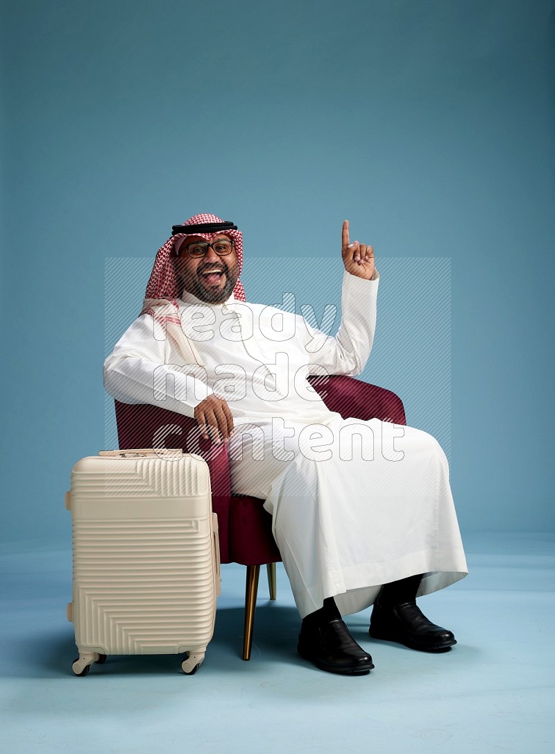 Saudi Man with shimag sitting on chair with a travel luggage on blue background