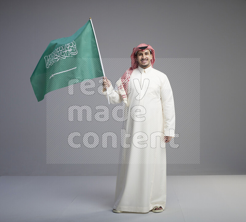 A Saudi man standing wearing thob and red shomag raising big Saudi flag on gray background