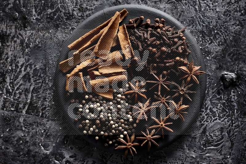 Cinnamon sticks, cloves, star anise and black and white peppers on a black plate on textured black background