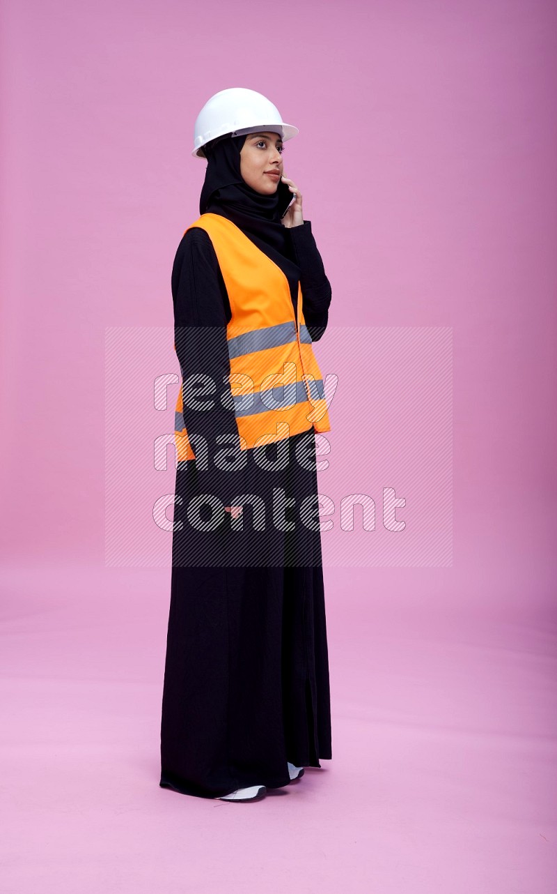 Saudi woman wearing Abaya with engineer vest and helmet standing talking on phone on pink background
