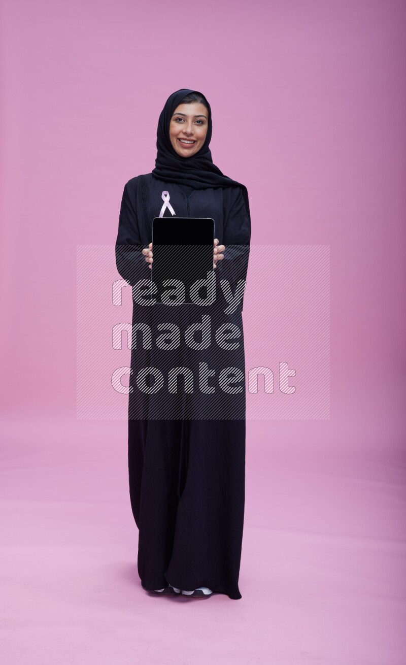 امرأة سعودية ترتدي شريطة زهرية على العباية تقف وتعرض الجهاز اللوحي على خلفية زهرية