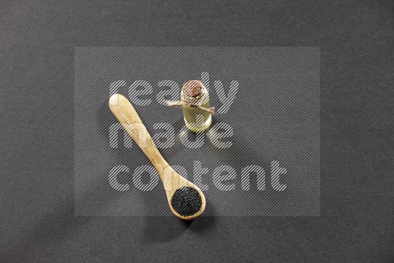 A wooden spoon full of black seeds and a glass bottle of black seeds oil on a black flooring
