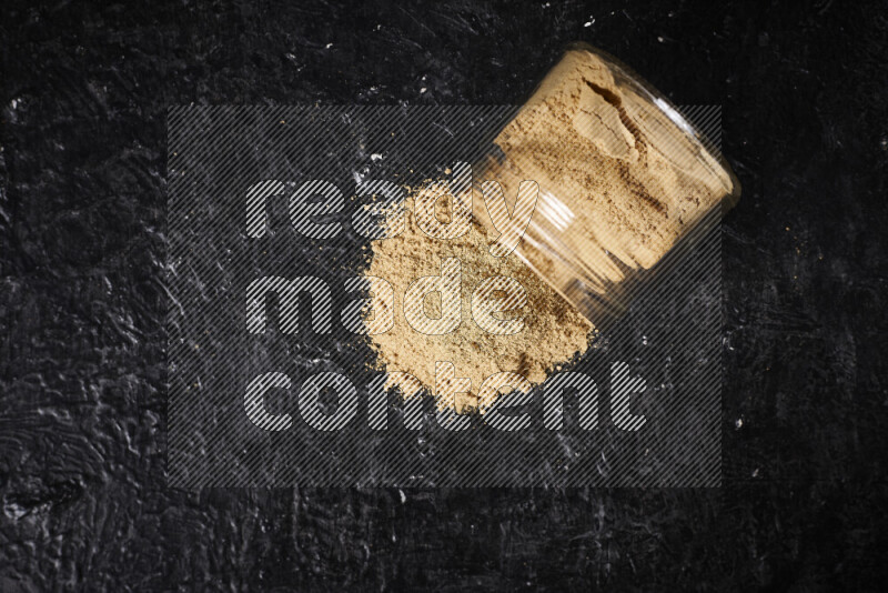 A glass jar full of ground ginger powder flipped with some spilling powder on black background