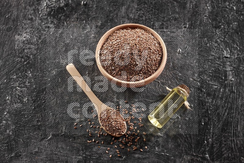 A wooden bowl and spoon full of flaxseeds and a glass bottle of flaxseeds oil on a textured black flooring