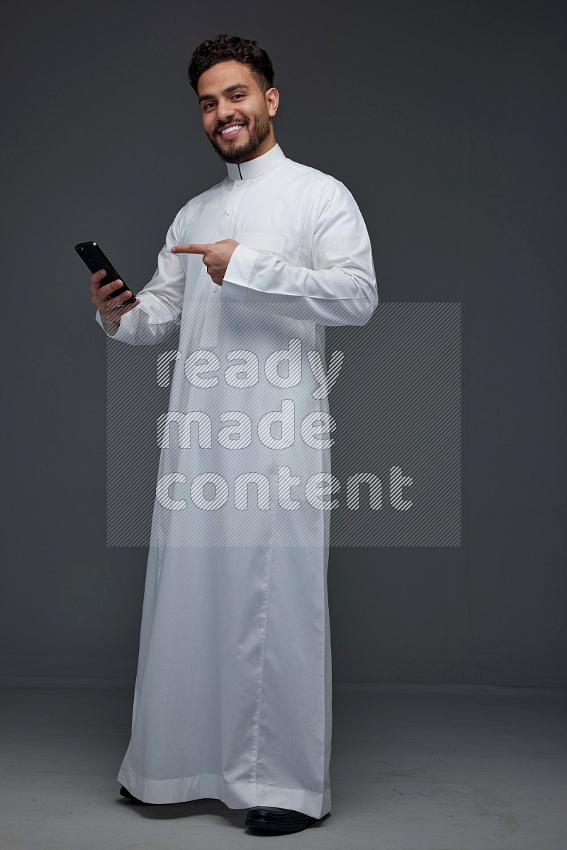 A Saudi man wearing Thobe and holding his phone while pointing to it eye level on a gray background