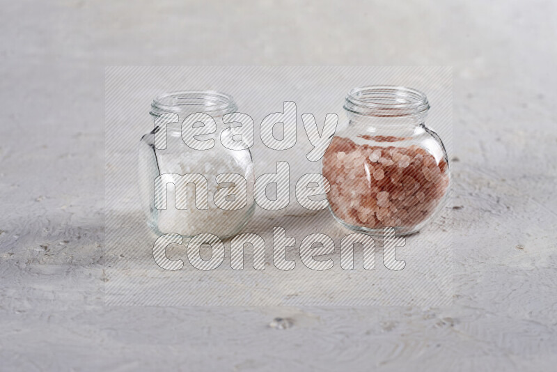 2 glass jars one is filled with coarse himalayan salt and the other with coarse sea salt on white background