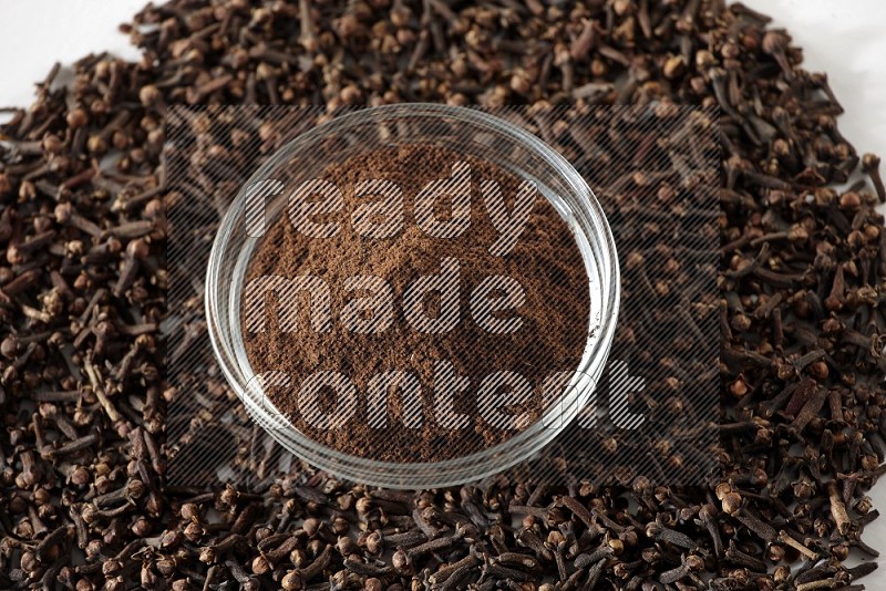 A glass bowl full of cloves powder and cloves grains spread on white flooring