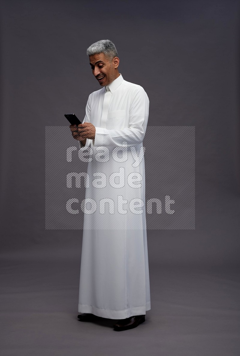 Saudi man wearing thob standing texting on phone on gray background