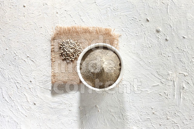 وعاء فخاري أبيض ممتلئ ببودرة الفلفل الأبيض موضوع على قطعة من الخيش مع حبوب الفلفل ومطحنة خشبية على أرضية بيضاء