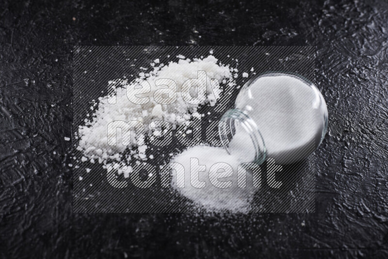 A glass jar full of table salt with some sea salt crystals beside it on a black background