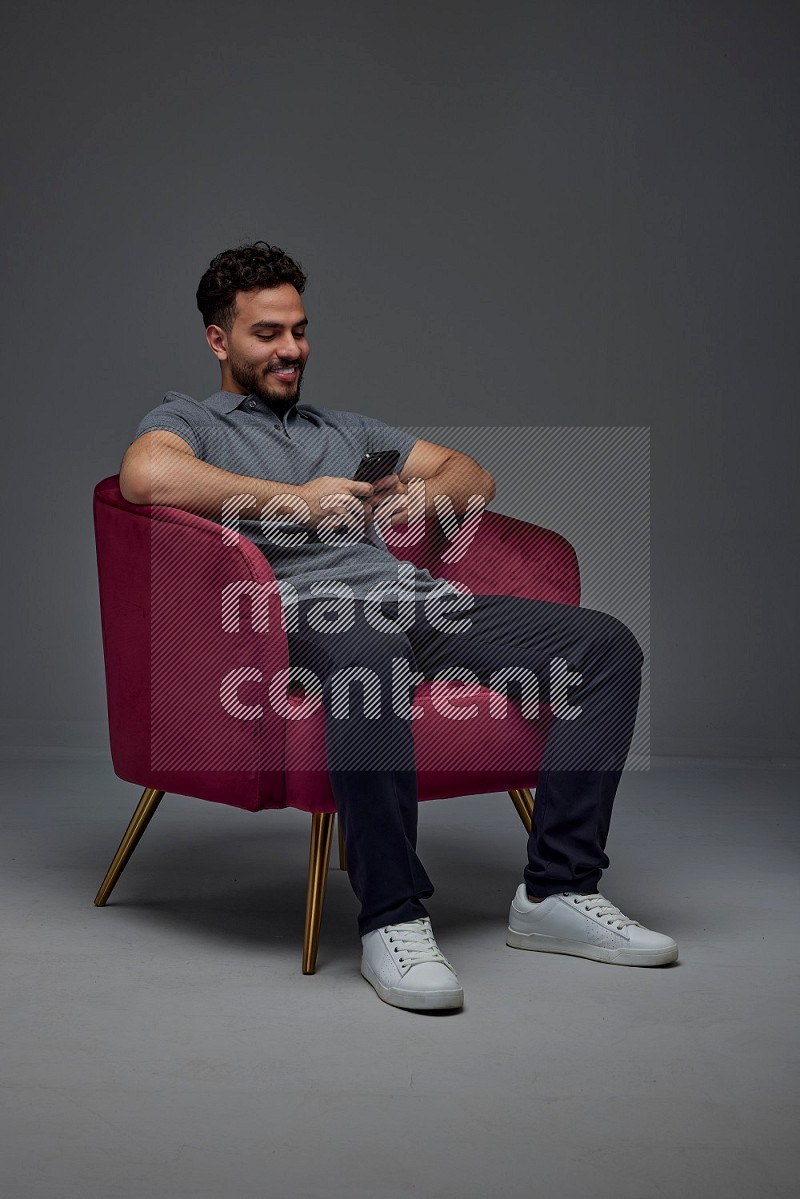 A man wearing casual and using his phone while sitting on a burgundy chair eye level on a gray background
