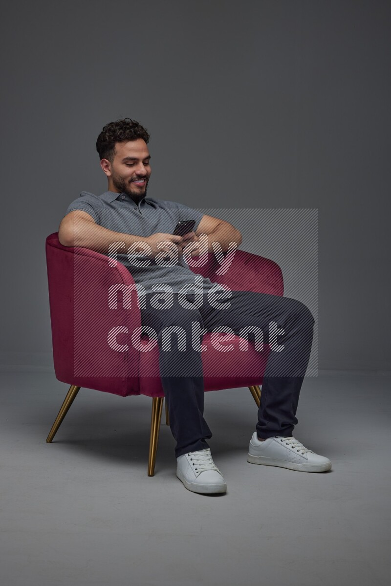A man wearing casual and using his phone while sitting on a burgundy chair eye level on a gray background