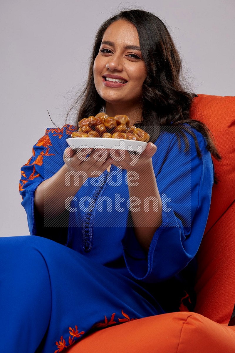 A Woman sitting on an orange beanbag wearing Jalabeya holding a plate of dates