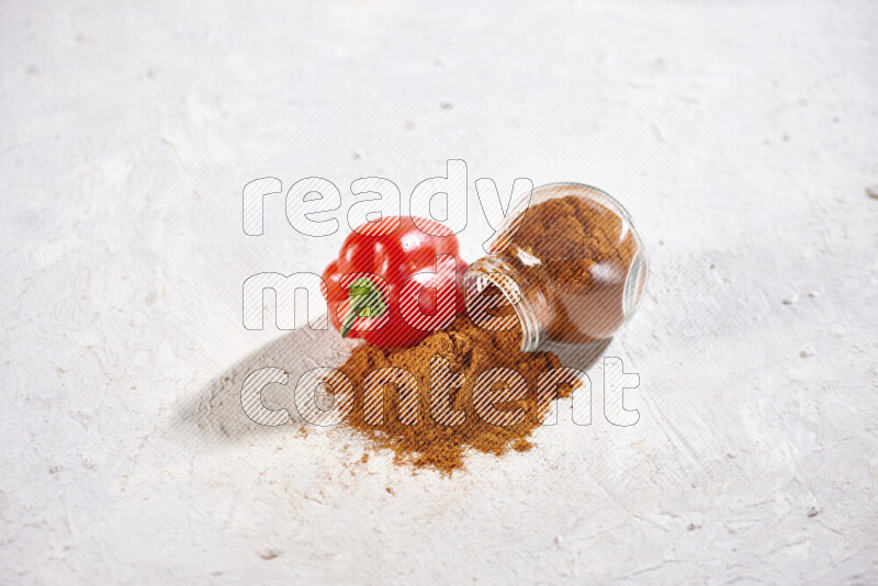 A glass jar full of ground paprika powder flipped with some spilling powder on white background