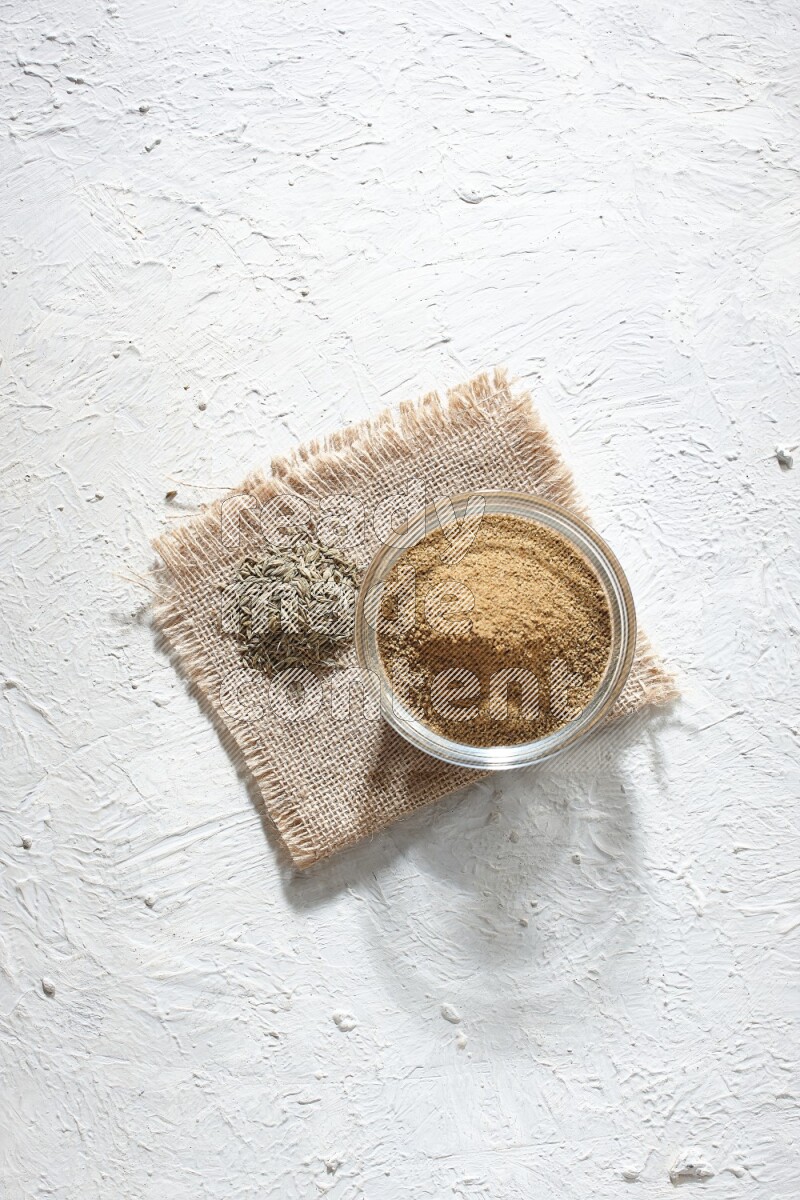 A glass bowl full of cumin powder with some of cumin seeds on burlap piece on a textured white flooring