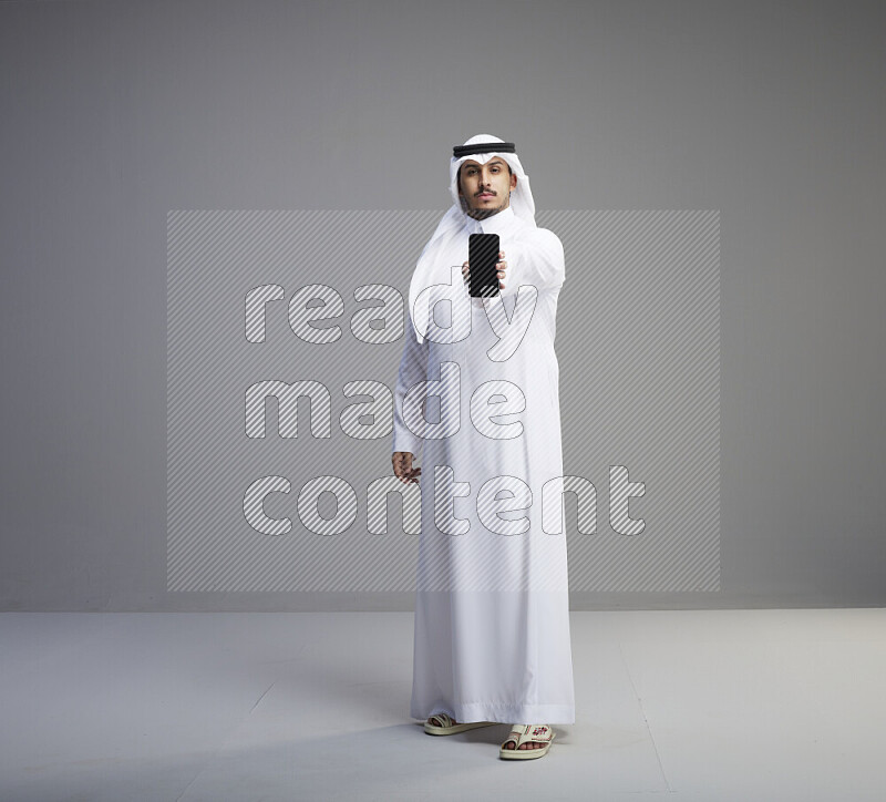 A Saudi man standing wearing thob and white shomag showing phone to camera on gray background