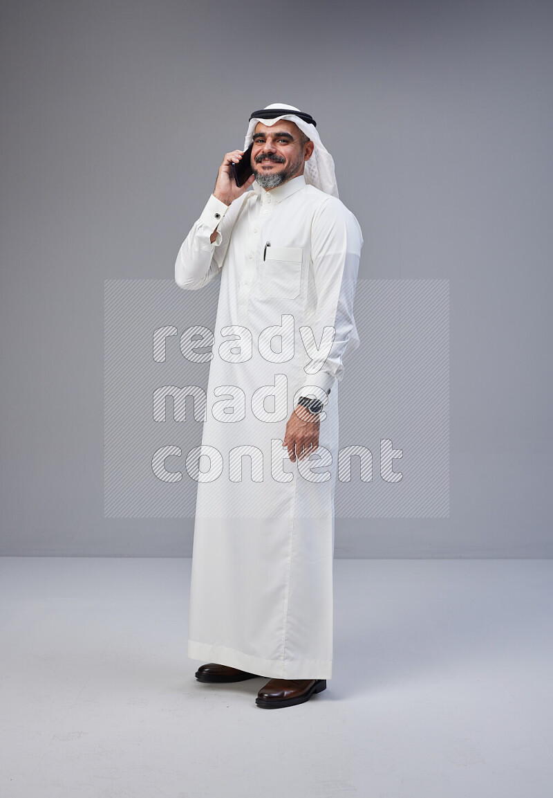 Saudi man Wearing Thob and white Shomag standing talking on phone on Gray background