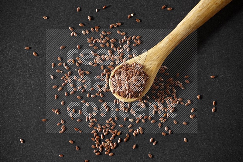 A wooden spoon full of flaxseeds and seeds spread beside it on a black flooring