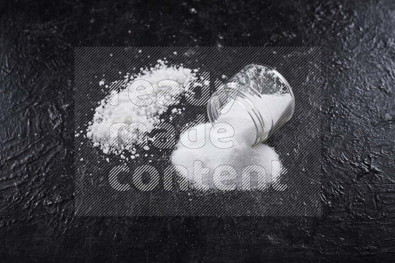 A glass jar full of table salt with some sea salt crystals beside it on a black background