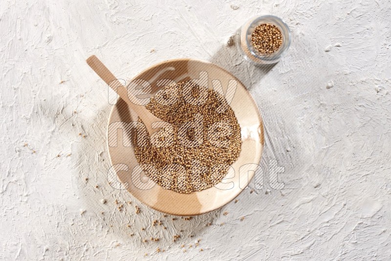 A beige pottery plate full of mustard seeds and a wooden spoon in it with a glass jar filled with the seeds on a textured white flooring
