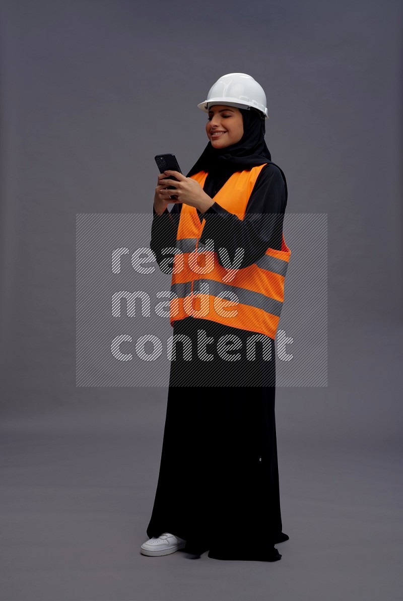 Saudi woman wearing Abaya with engineer vest standing texting on phone on gray background