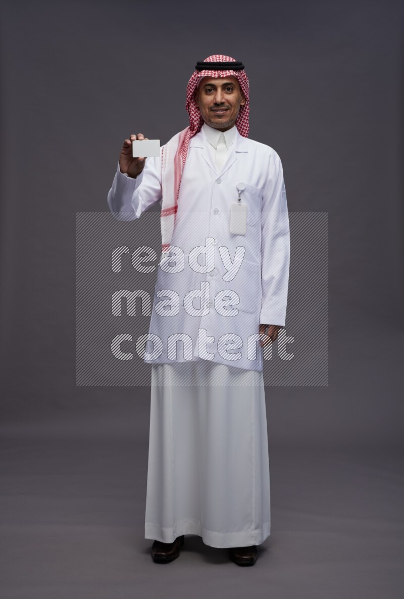 Saudi man wearing thob with lab coat and shomag with pocket employee badge standing holding ATM card on gray background