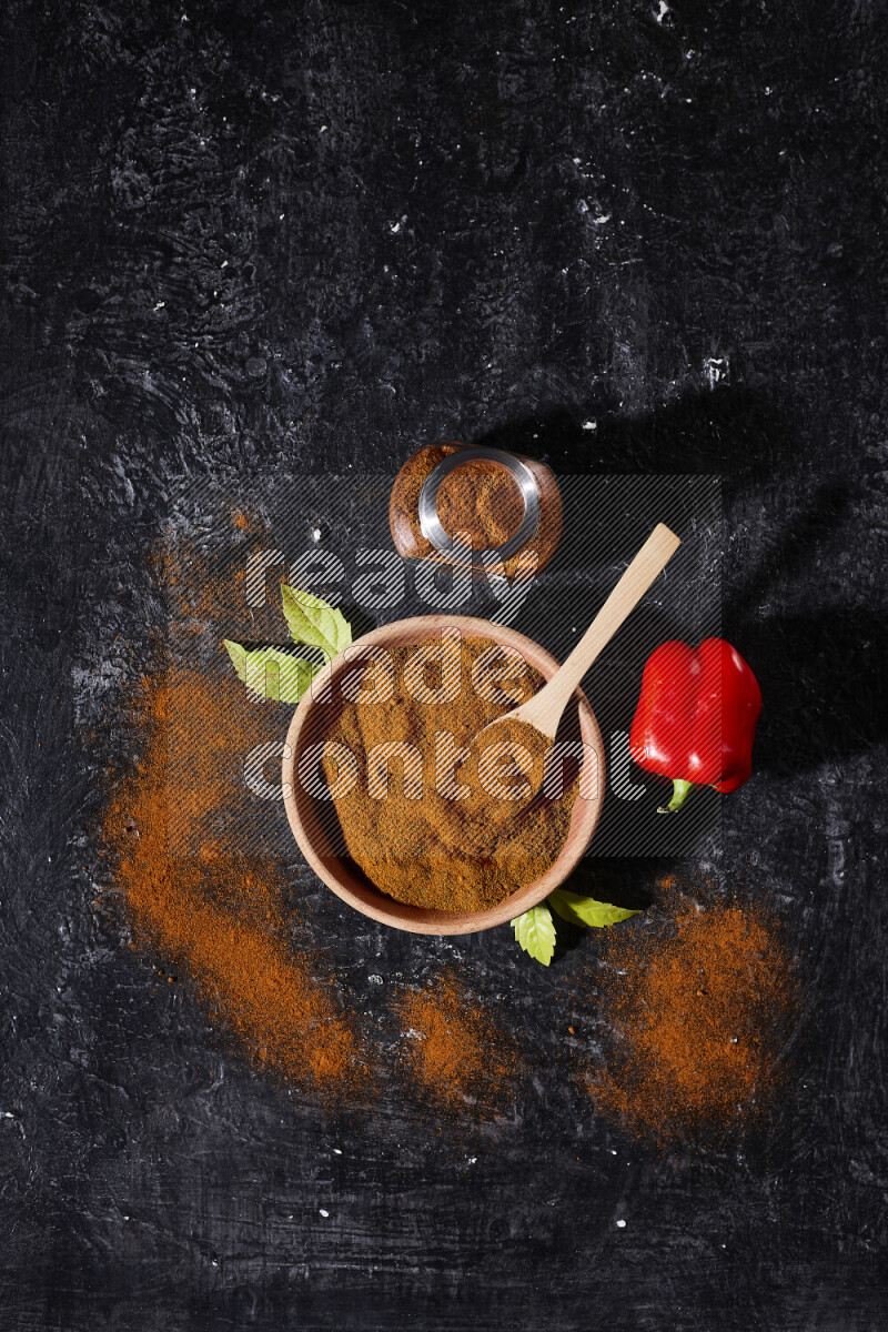 A wooden bowl full of ground paprika powder with a glass jar beside it and a red bell pepper on black background