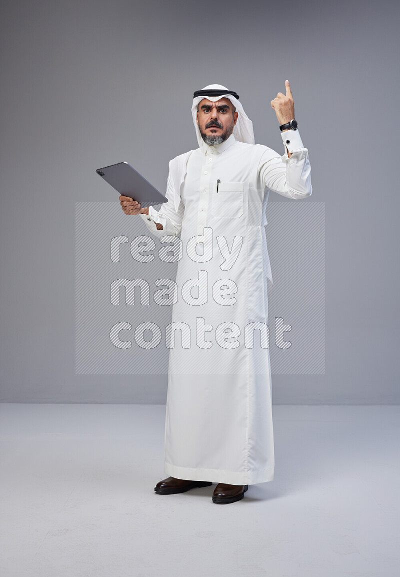 Saudi man Wearing Thob and white Shomag standing working on tablet on Gray background