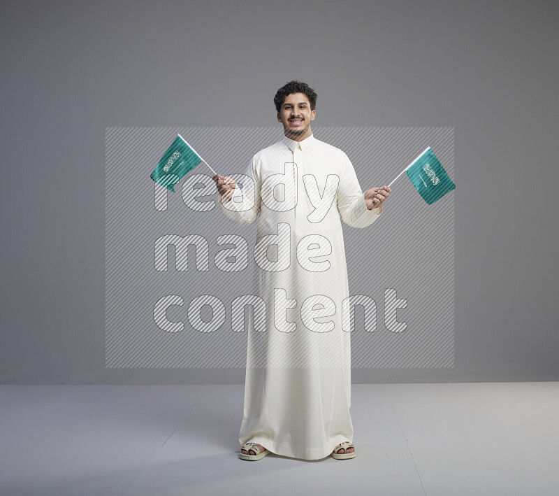 رجل سعودي يرتدي ثوب ابيض يحمل علم السعودية على خلفية رمادي