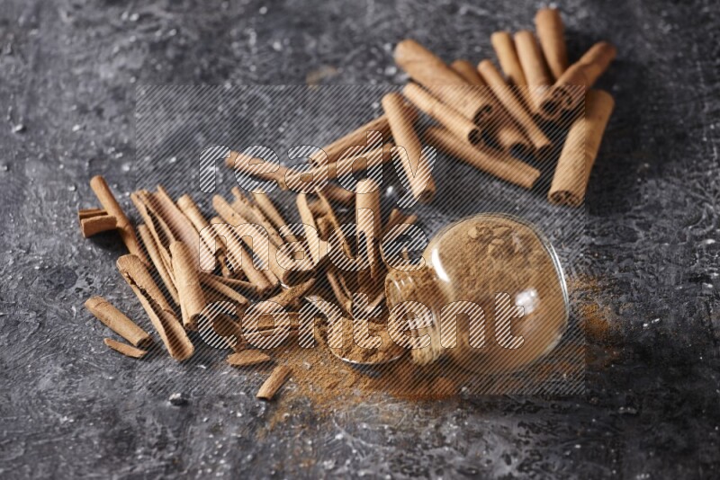 Herbal glass jar full cinnamon powder flipped and a metal spoon full of powder surrounded by cinnamon sticks on textured black background in different angles