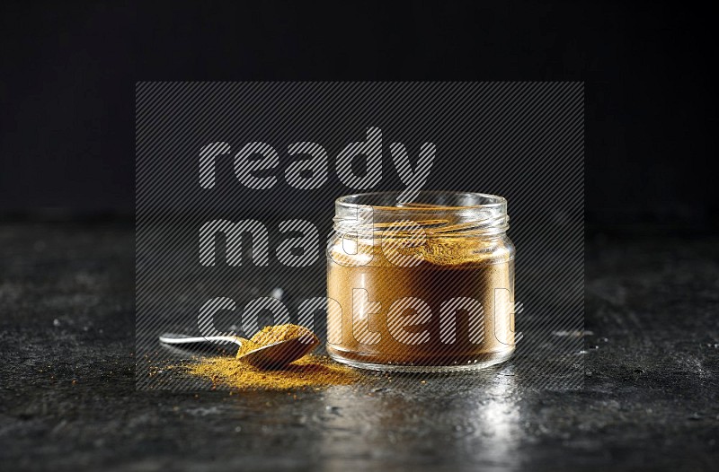 A glass jar and a metal spoon full of turmeric powder on a textured black flooring