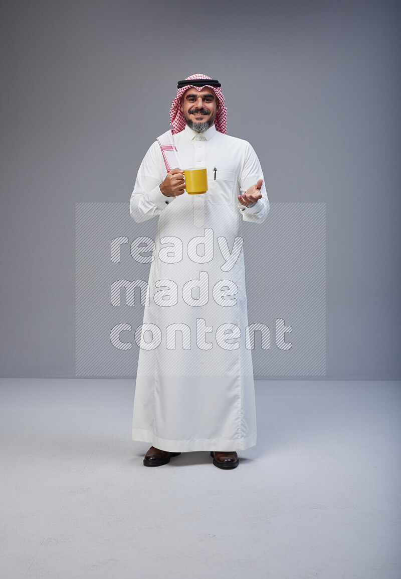 Saudi man Wearing Thob and red Shomag standing holding mug on Gray background